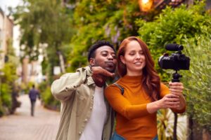 Young Couple Travelling Through City Together Vlogging To Video Camera On Handheld Tripod